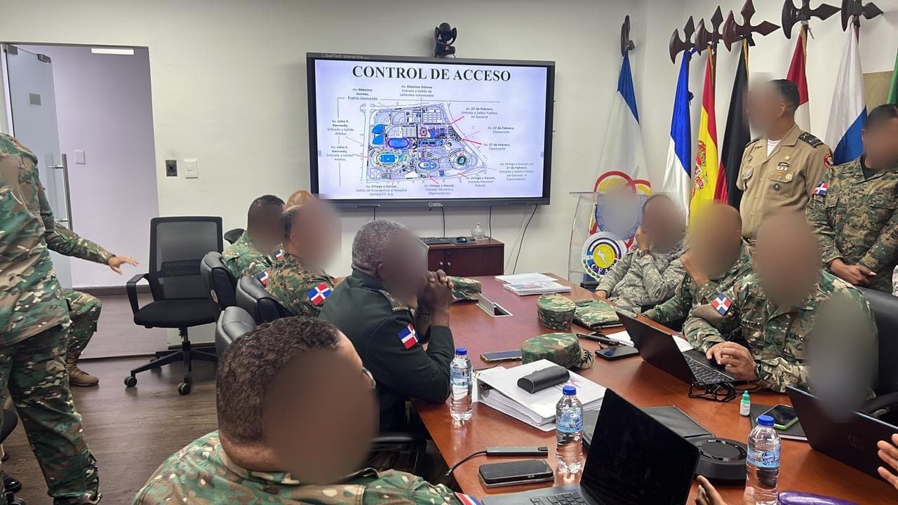 Reunión de planificación del control de acceso ante el plano del recinto en pantalla, con los equipos de seguridad de los Juegos 2026