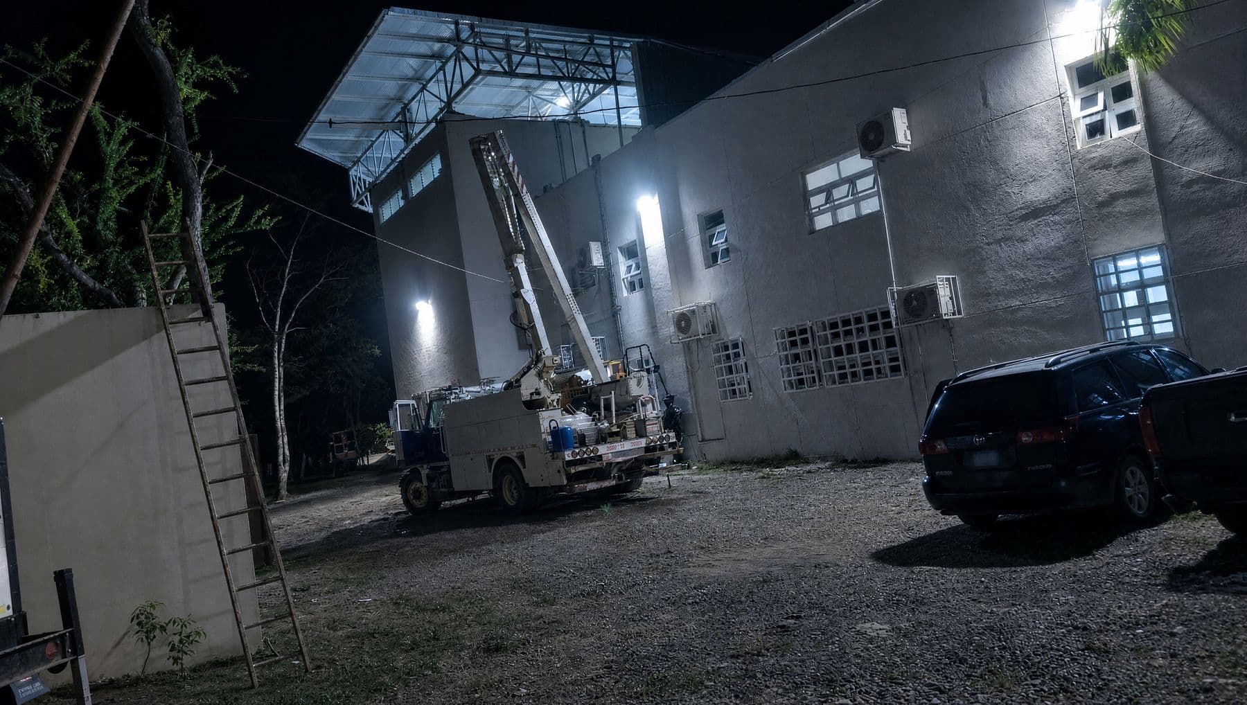 Camión con grúa de canasta durante el montaje nocturno de equipos en la fachada de un pabellón de los Juegos Centroamericanos y del Caribe 2026