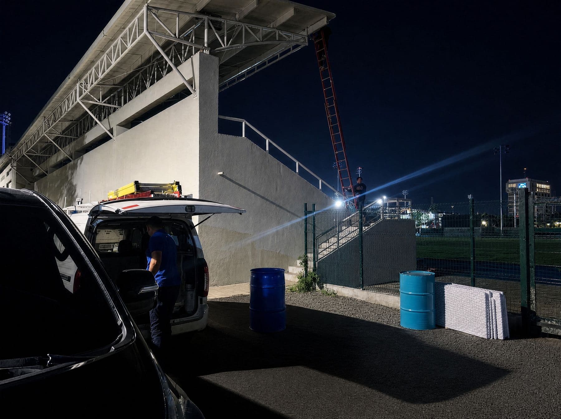 Montaje nocturno junto al graderío: vehículo de trabajo y escalera contra la torre de iluminación