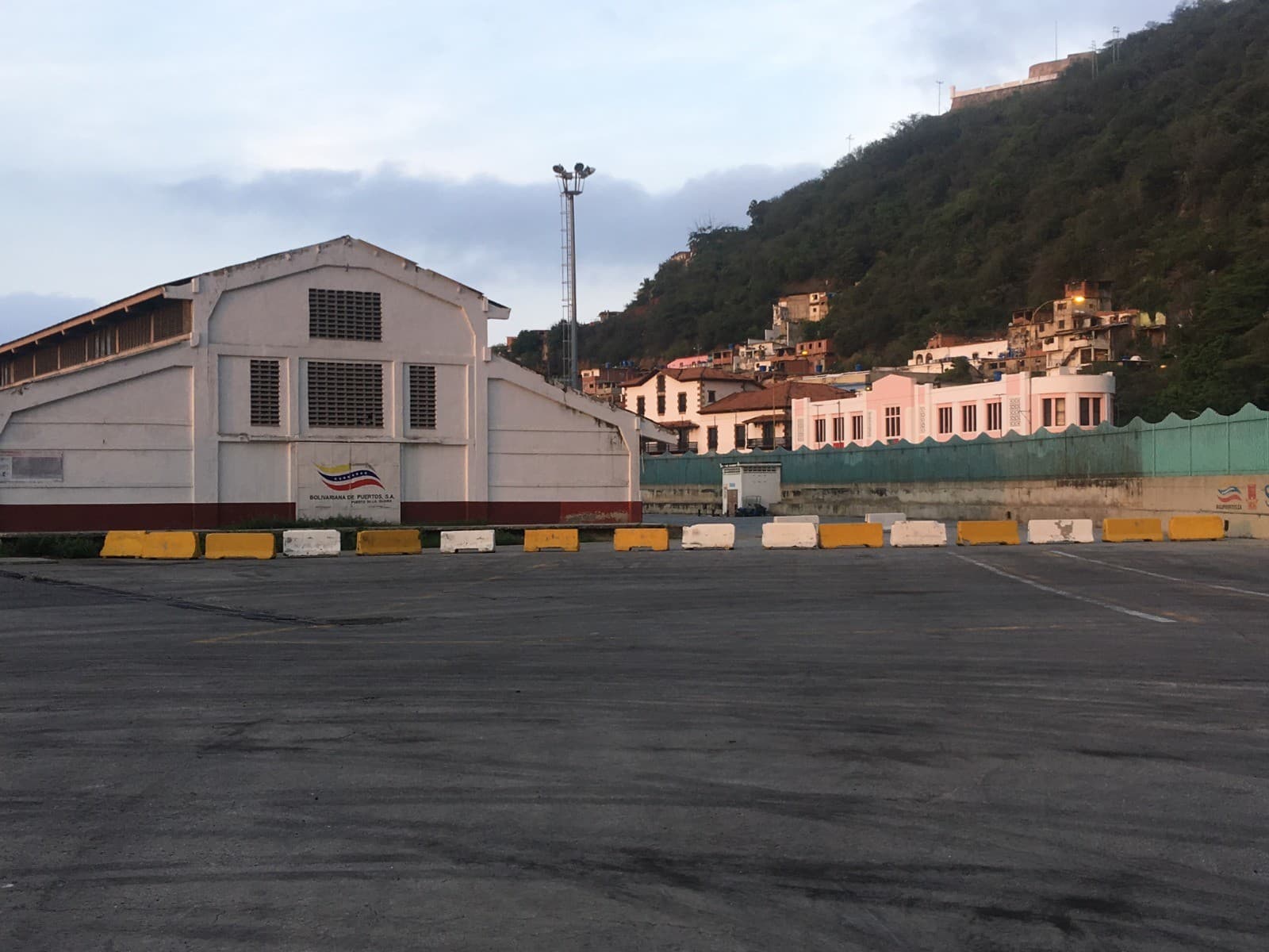 Explanada del muelle de La Guaira con barreras de hormigón, galpón portuario blanco y cerro con viviendas al fondo